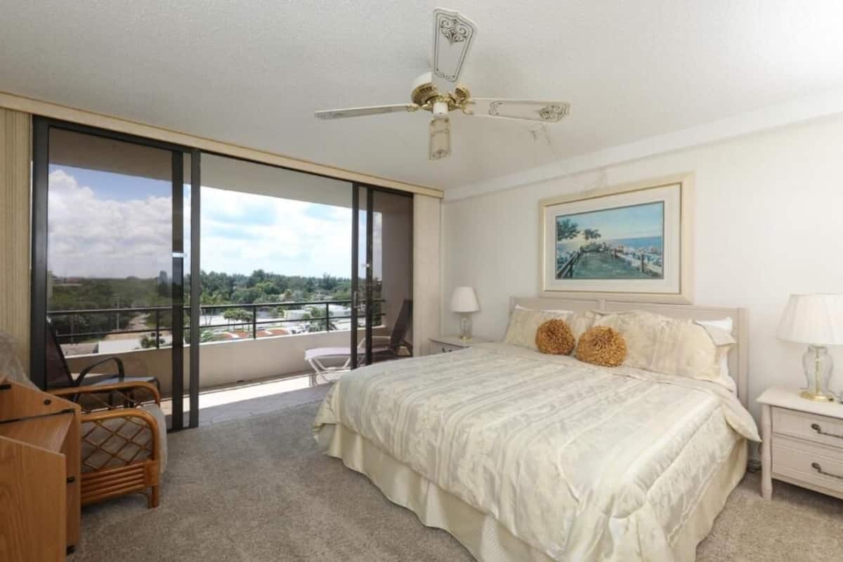 A hotel room with a large bed, a balcony with a view, a ceiling fan, and a painting on the wall.