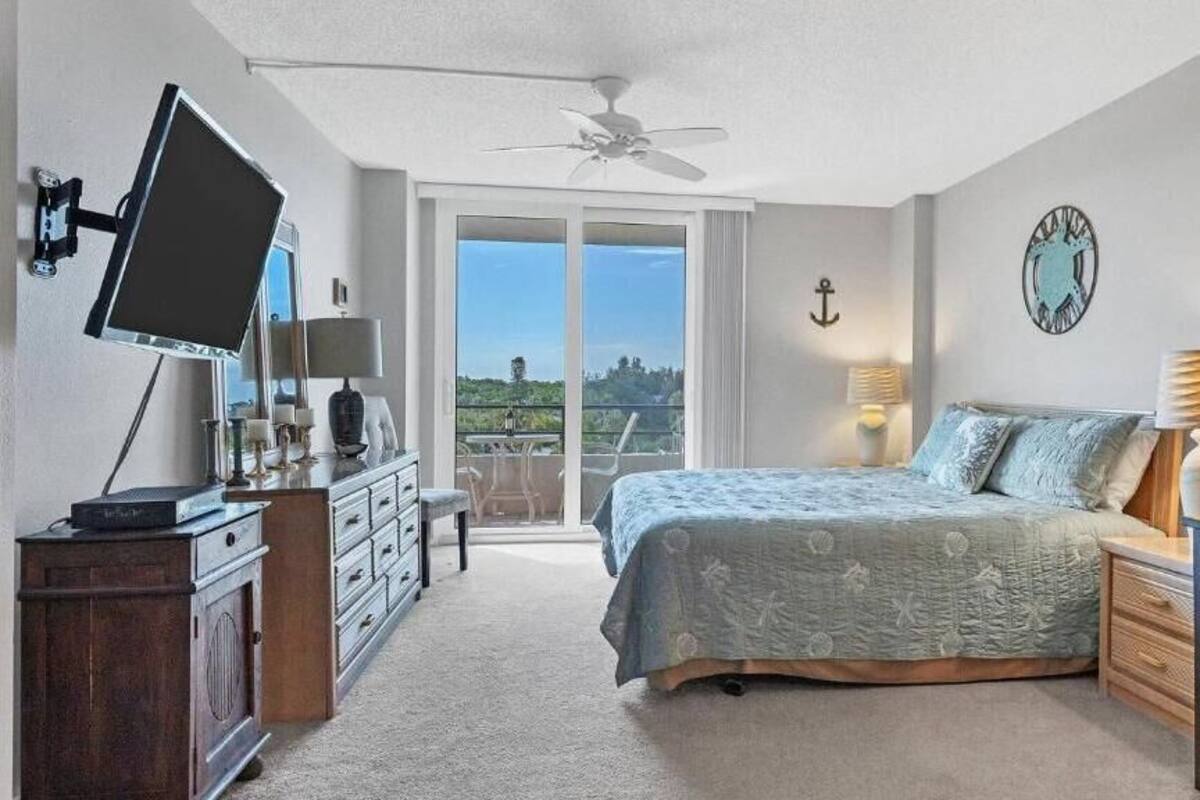 A bedroom with a bed, a desk, a chair, a TV, a dresser, and a balcony with a view.