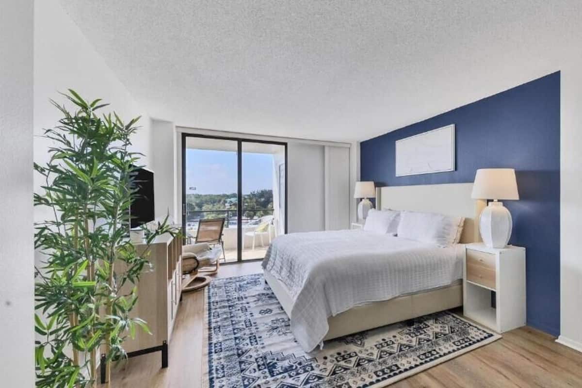 A bedroom with a bed, bedside tables, a lamp, a plant, and a view of the outdoors.