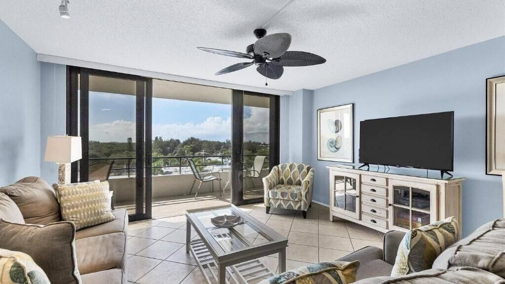 A spacious living room with a large flat-screen TV, comfortable seating, a ceiling fan, and a balcony with a scenic view.
