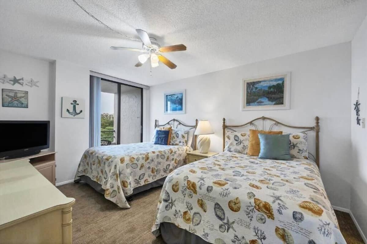 A bedroom with two beds, a ceiling fan, a TV, and a window with a view of greenery.