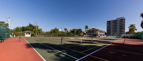 Una cancha de tenis al aire libre con una red, rodeada por una valla, y contigua a un edificio.