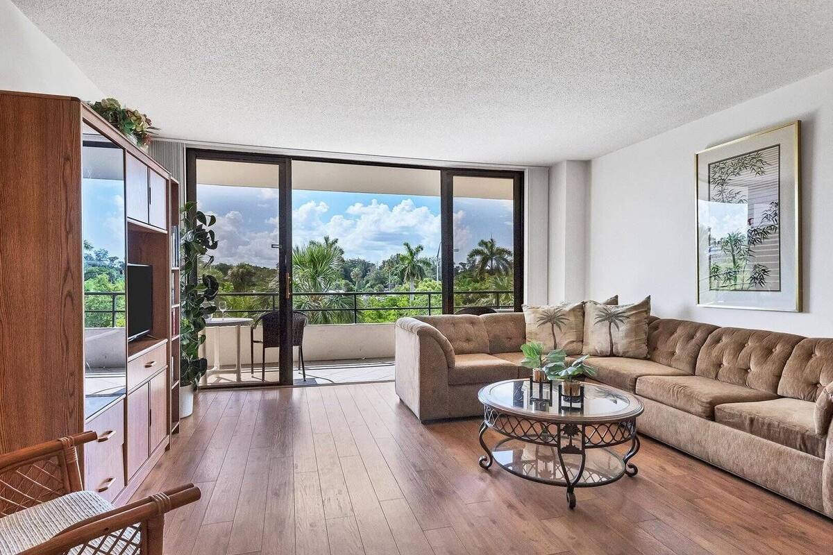 A spacious living room with a large sliding glass door leading to a balcony, a brown sofa, a glass coffee table, and a wooden cabinet.