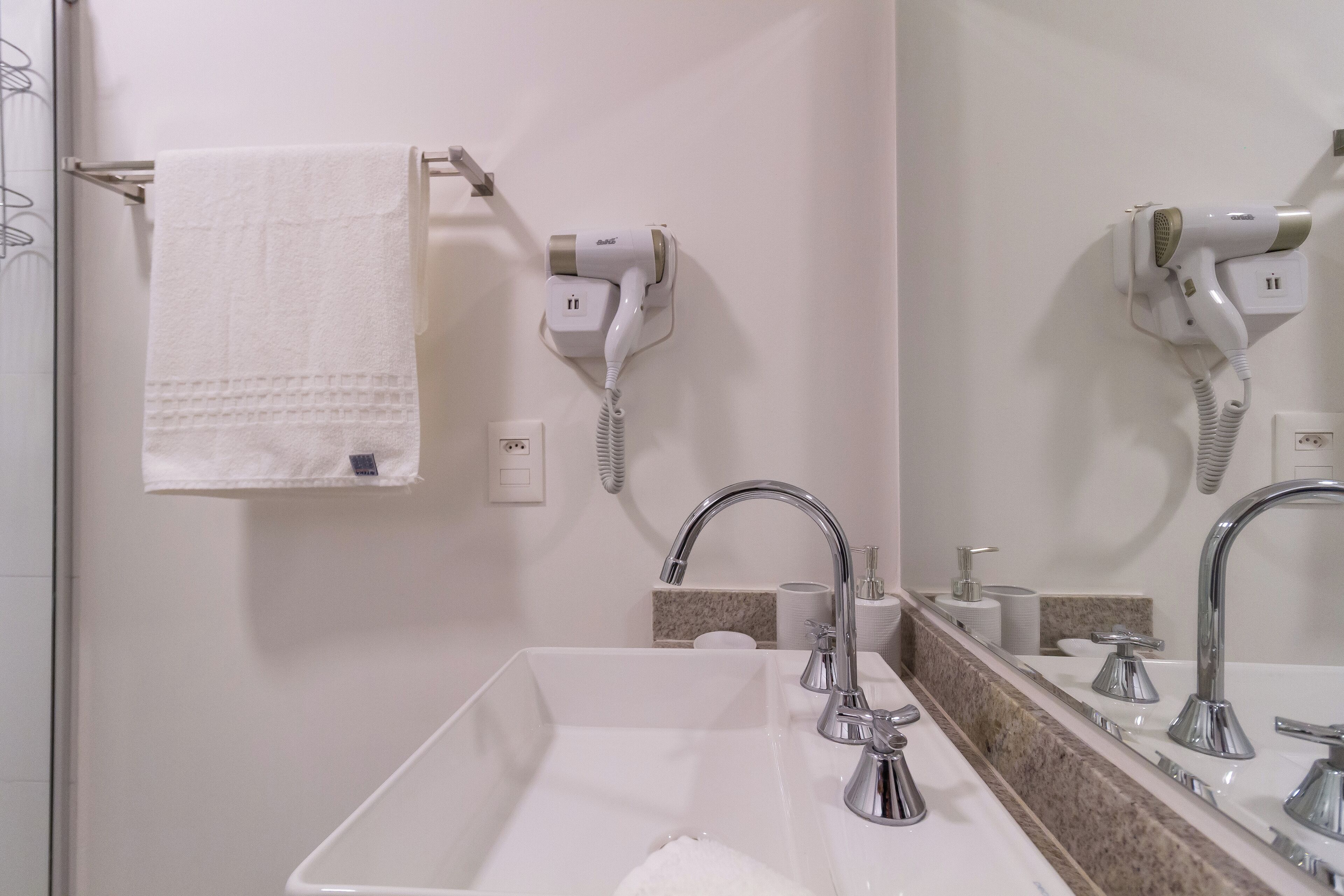 Une salle de bain avec un lavabo blanc, deux robinets, un porte-serviettes avec une serviette blanche, et deux sèche-cheveux fixés au mur.