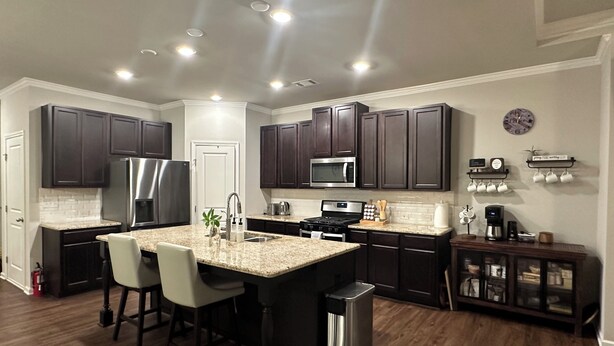 A modern kitchen with dark wood cabinets, stainless steel appliances, and a central island with bar stools. - Model- Home Modern, Spacious, and Great Location (Buford)