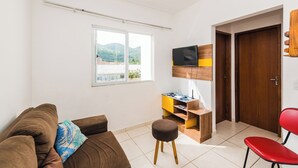 A living room with a brown sofa, a small stool, a wooden TV stand, and a red chair. - Cozy Apartment near Cambirela CFT (Palhoça)