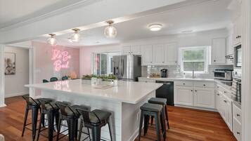A modern kitchen with a central island, bar stools, white cabinetry, stainless steel appliances, and a neon sign.