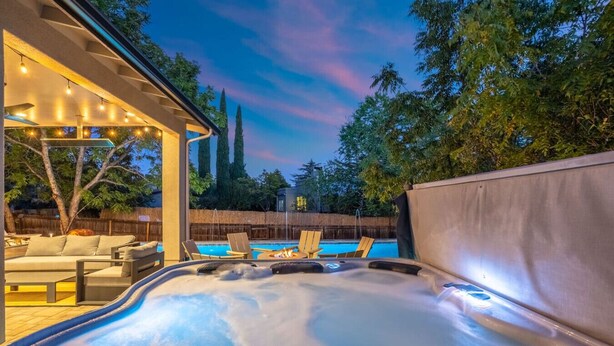 Un bain à remous avec vue sur une piscine et des chaises longues, entouré d’arbres et d’une clôture en bois.