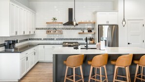 Une cuisine moderne avec des meubles blancs, un îlot central doté de tabourets, un réfrigérateur en acier inoxydable et un éclairage suspendu.