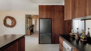 A modern kitchen with wooden cabinets, a black refrigerator, and a dishwasher. There is a decorative wall piece and a woven basket on the wall. - Oceanfront Escape – Two-Level Penthouse with Private Rooftop & Pool (Tulum)