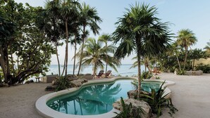 A private pool with a curved edge, surrounded by palm trees and rocks, overlooking a beach. - Oceanfront Escape – Two-Level Penthouse with Private Rooftop & Pool (Tulum)
