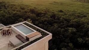 A rooftop terrace with a rectangular pool, wooden furniture, and a view of a dense forest.
