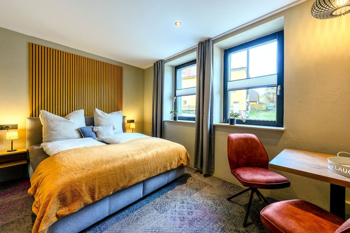 A hotel room with a large bed, a desk, and a chair. There is a window with curtains and a view of the outside.