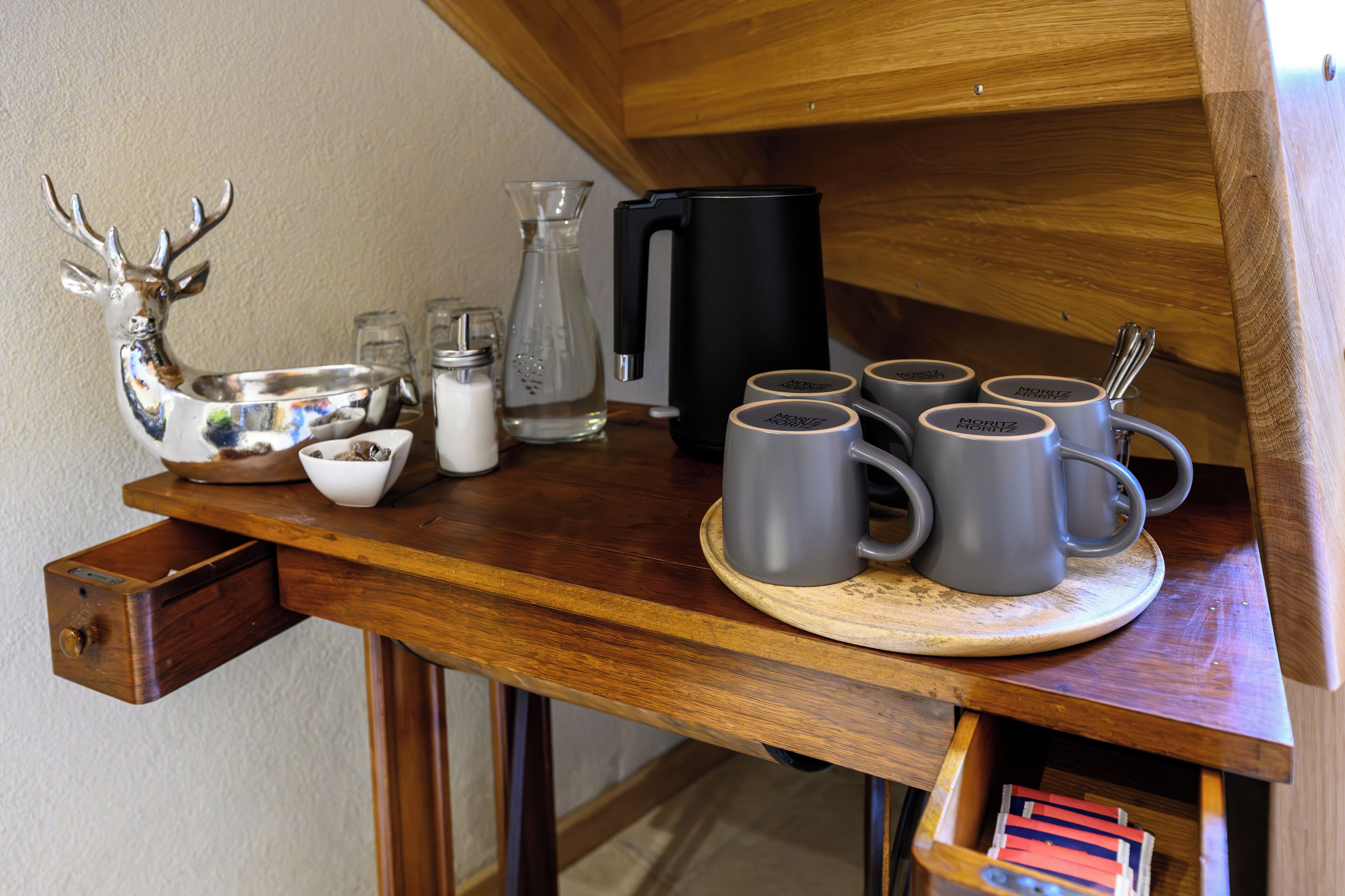 Una mesa de madera con una cafetera, una jarra de vidrio, una figurita de ciervo de cerámica, un tazón blanco y una bandeja con seis tazas grises.