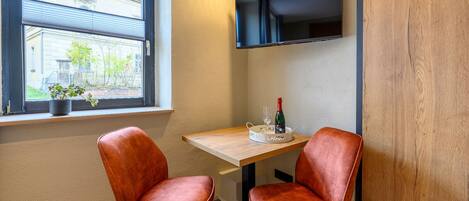 Habitación de esquina con una mesa de madera, dos sillones de cuero, un televisor montado en la pared y una ventana con vistas a un edificio y vegetación.