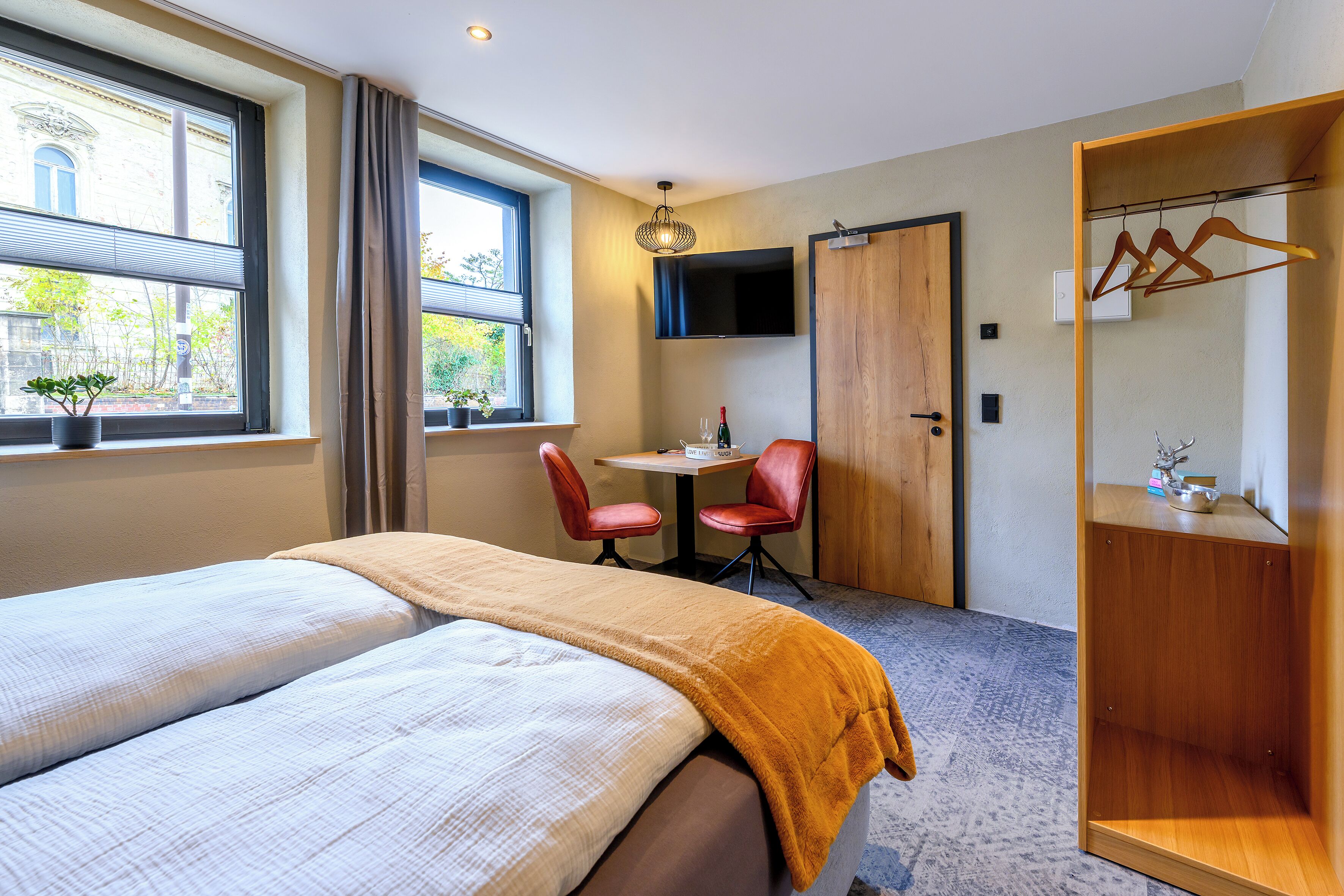 A hotel room with a bed, a desk, two chairs, a TV, a wardrobe, and a window with a view of a building.