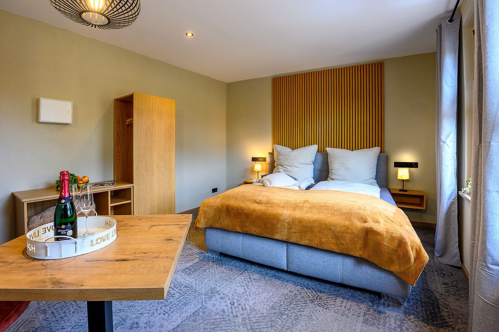 A hotel room with a bed, bedside tables, a wooden nightstand, a bottle of champagne, and a tray with glasses.
