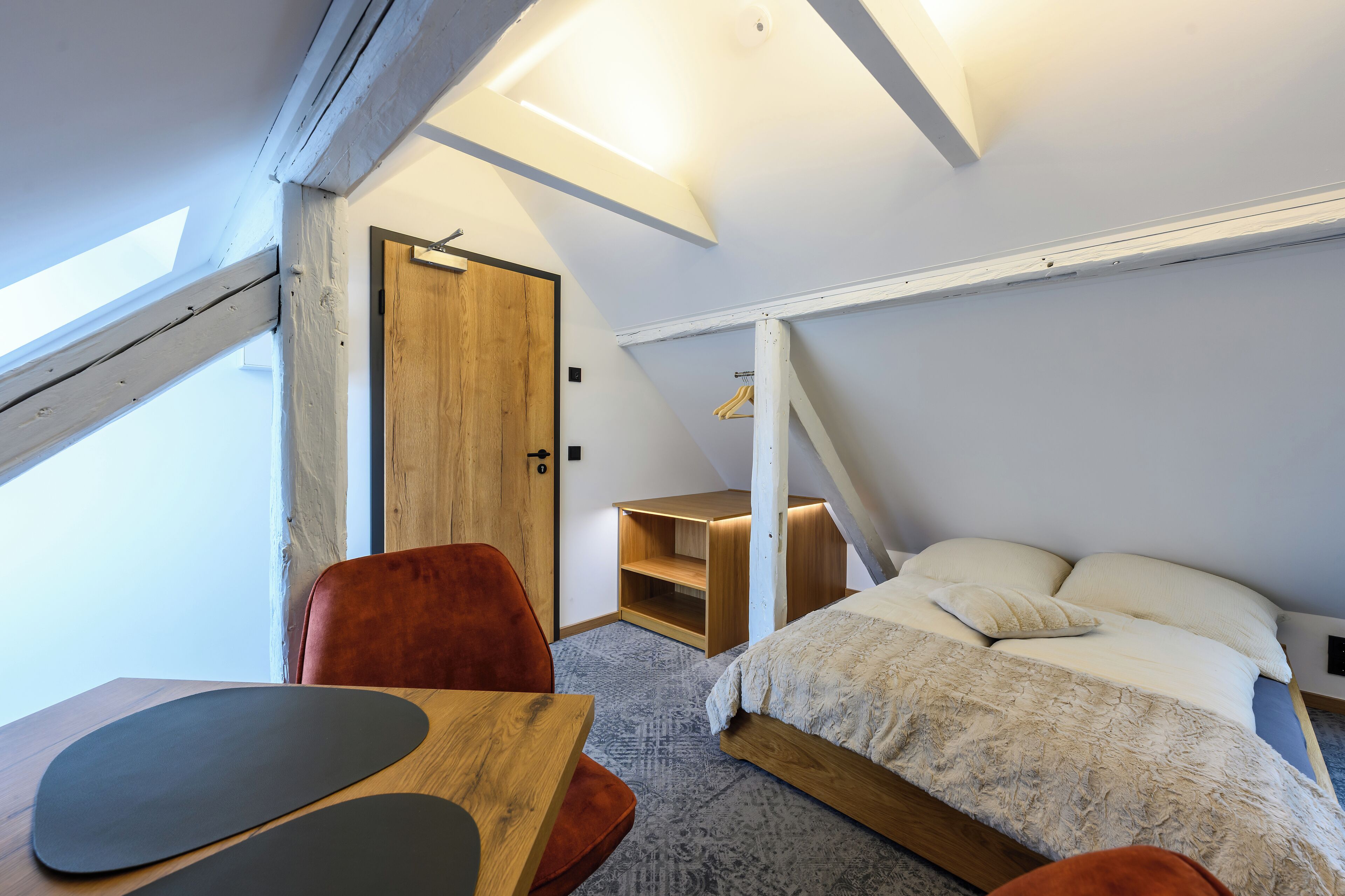 Un dormitorio con techo inclinado, cama con ropa blanca, mesita de noche de madera, una silla roja y una pequeña mesa con un tapete negro.