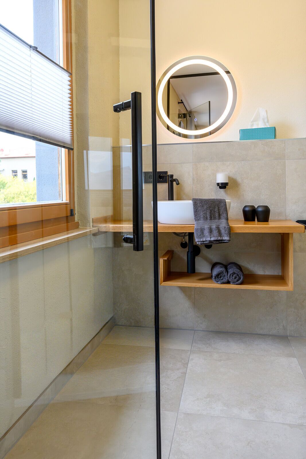 Un baño moderno con cabina de ducha de vidrio, un espejo redondo con iluminación integrada, un lavabo blanco y un estante de madera con toallas.