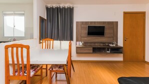 A modern living room with a white dining table, two wooden chairs, a flat-screen TV mounted on a wooden panel, a small cabinet below the TV, and a wooden door. - Cozy and practical apartment in a residential neighborhood (Palhoça)