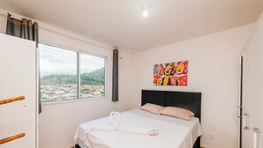A bedroom with a bed, a window with a view, a painting on the wall, and a door. - Cozy and practical apartment in a residential neighborhood (Palhoça)