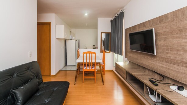 A compact living space with a black leather sofa, a wooden dining table with two chairs, a kitchen area with a white refrigerator, and a mounted flat-screen TV on a wooden wall. - Cozy and practical apartment in a residential neighborhood (Palhoça)