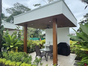 Un espace extérieur aménagé en patio, doté d’un coin salon couvert, d’un barbecue et d’une végétation luxuriante.