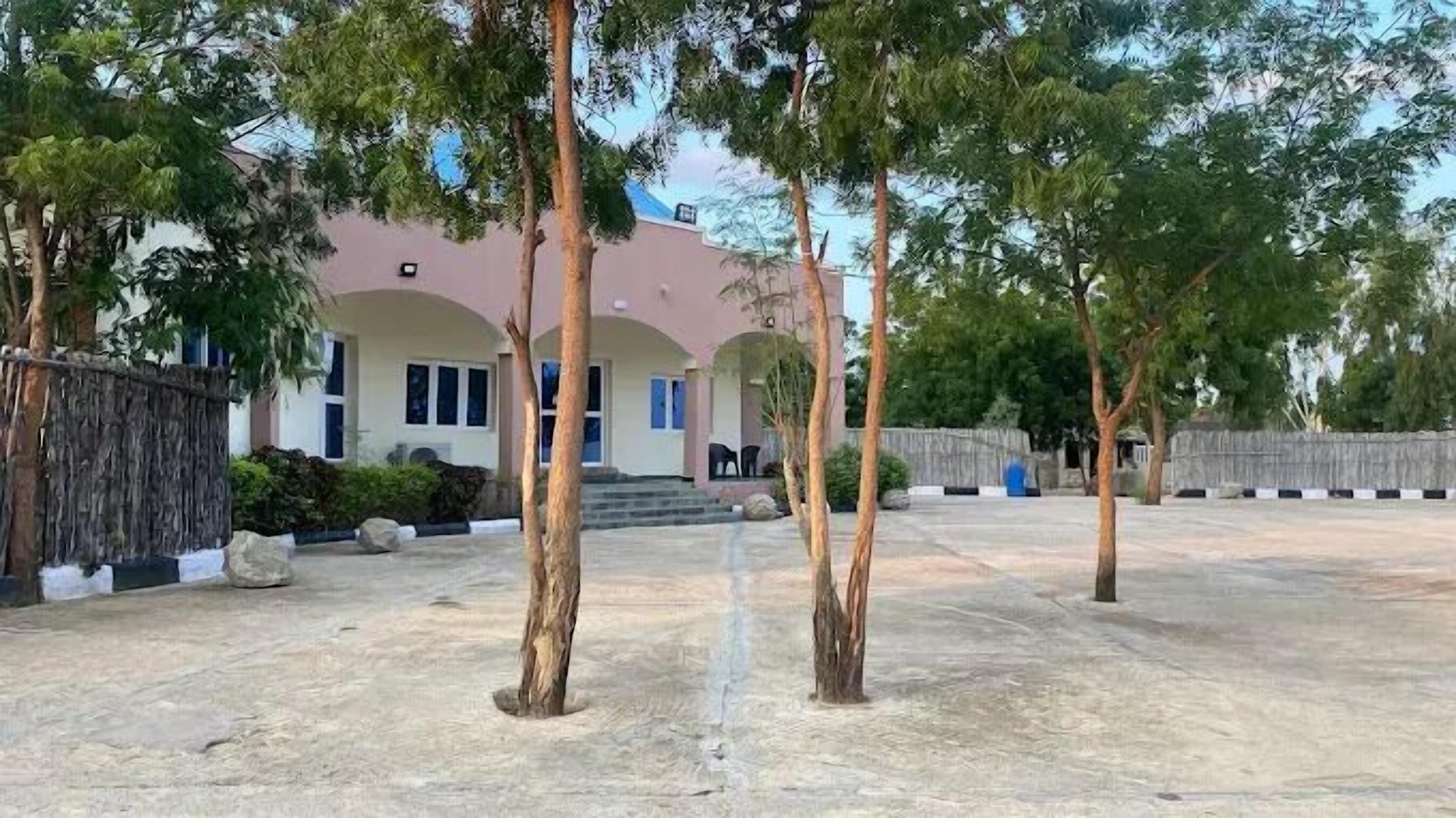 A building with a pink facade and arched windows, surrounded by trees and a sandy ground.