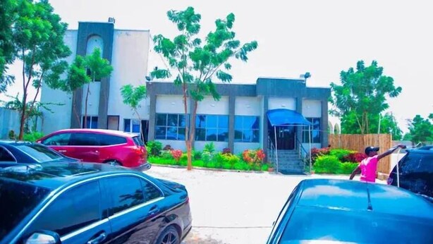 A parking lot with several cars, a building with a modern design, and a person in pink. - MTown Hotel and Suites  (Dutse)