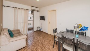 En stue med en beige sofa, et spisebord med stole, et fjernsyn ophængt på væggen og et vindue med gardiner.