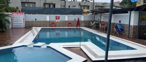 Piscina com placa de 'POOL RULES', placa 'NOVO RÓTULO' e área de estar coberta com cadeiras.