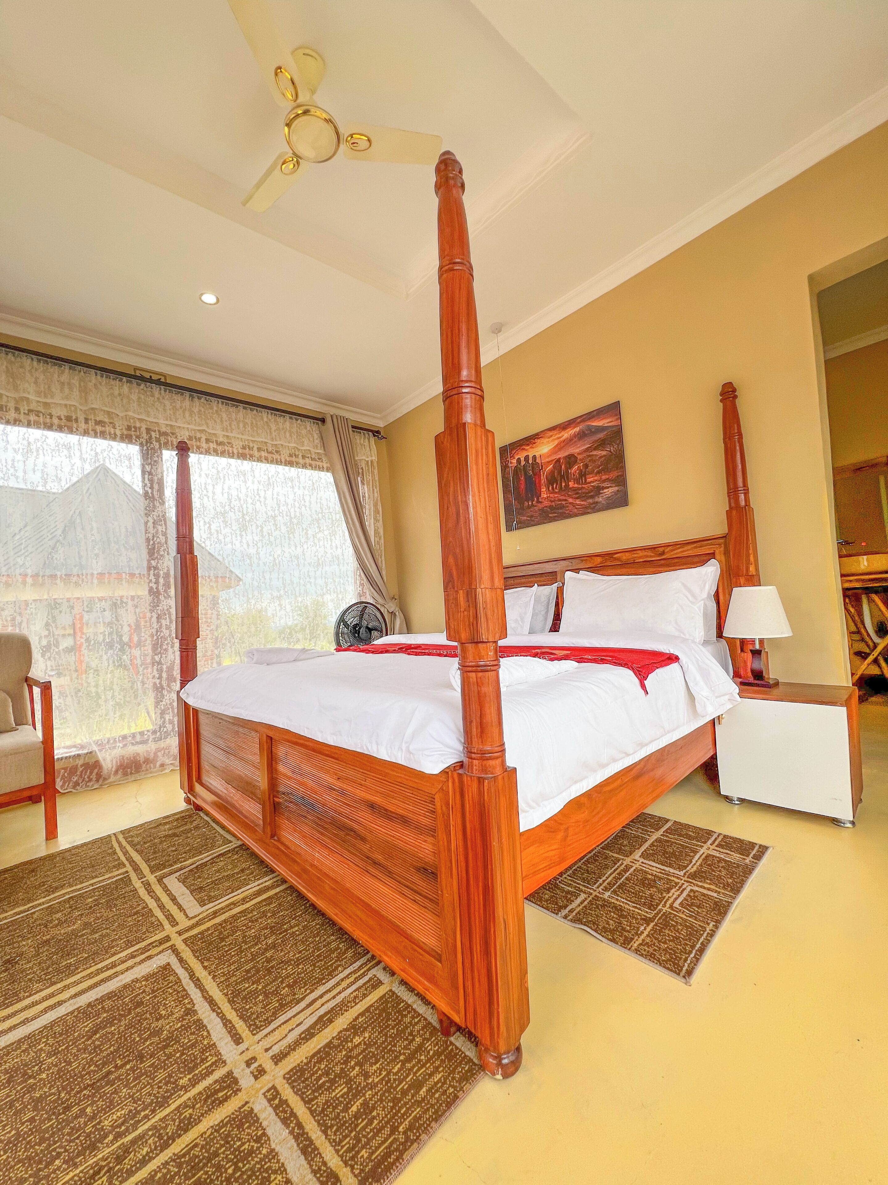 A four-poster bed with a wooden headboard, a bedside table with a lamp, a chair, and a large window with curtains.