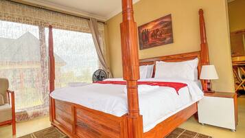 A four-poster bed with a wooden headboard, a bedside table with a lamp, a chair, and a large window with curtains.