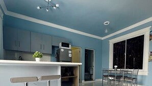 Une cuisine moderne avec des murs bleus, des plans de travail blancs et des appareils en acier inoxydable. On y trouve deux tabourets de bar et une table à manger avec des chaises.