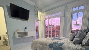 Une chambre avec un grand lit, un téléviseur fixé au mur, une fenêtre donnant sur une rangée de vélos et une petite étagère ornée d’objets décoratifs.