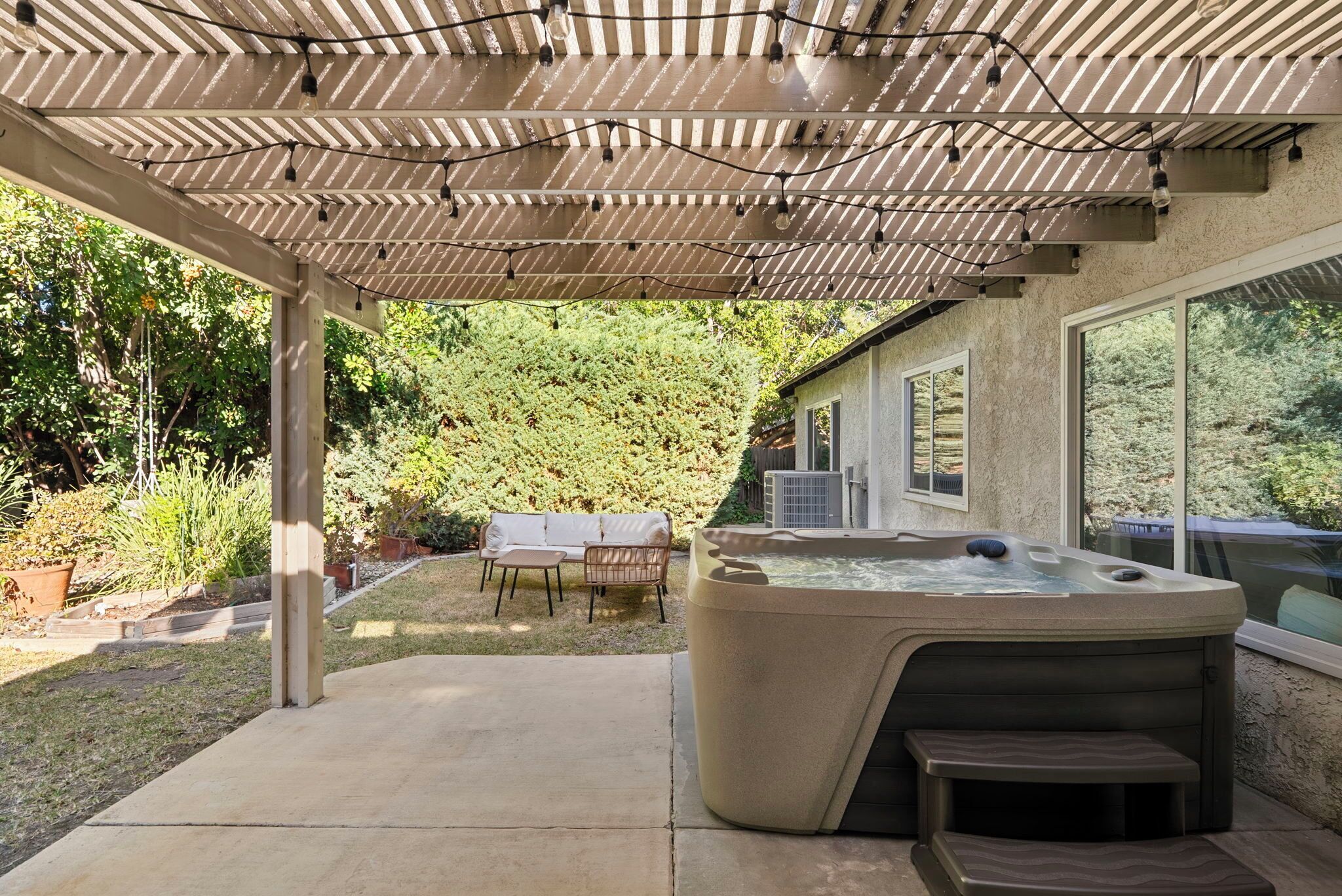 A hot tub under a pergola with string lights, a patio with outdoor furniture, and a view of a forested area.