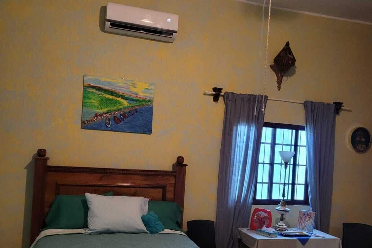 A bedroom with two beds, a painting on the wall, an air conditioning unit, a table with a lamp, and a window with curtains.