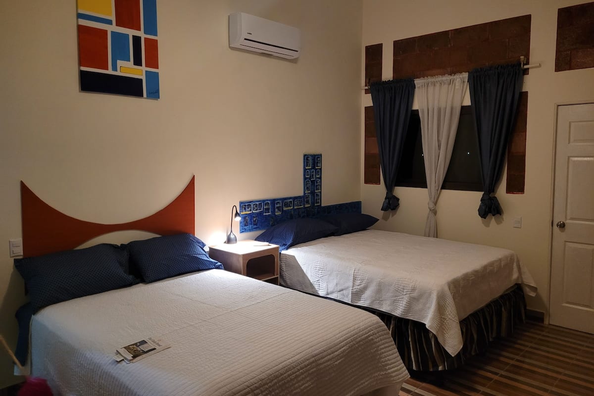 A hotel room with two beds, a nightstand, an air conditioning unit, a window with curtains, and a colorful abstract painting on the wall.