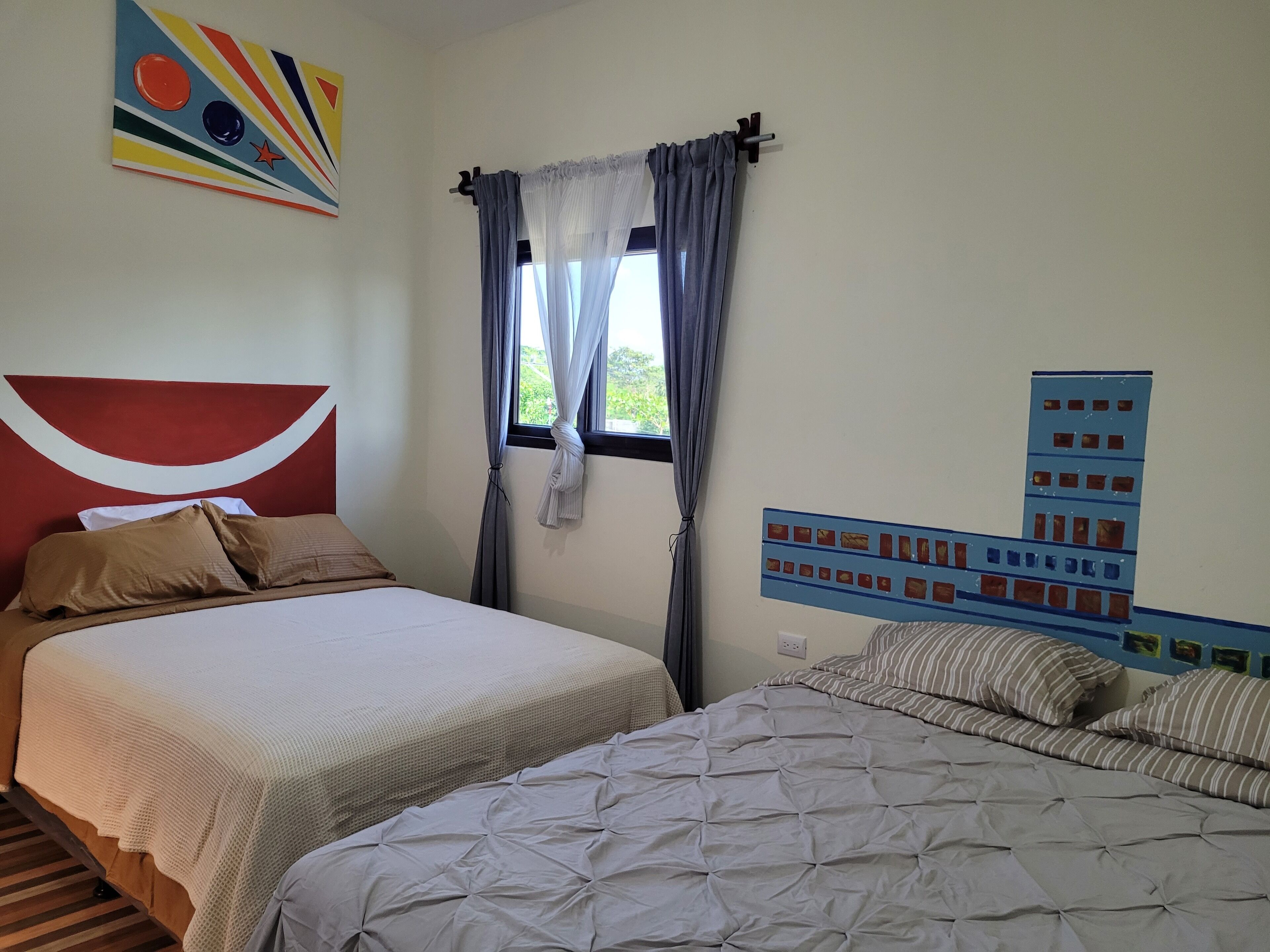 Een slaapkamer met twee bedden, een groot raam met gordijnen, een kunstwerk aan de muur en een lichtschakelaar op de muur.