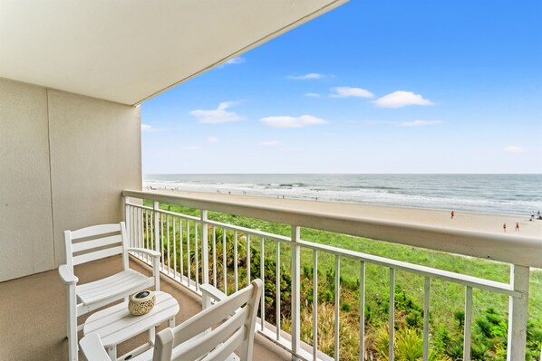 A balcony with white chairs and a small table, overlooking a beach with people and greenery.