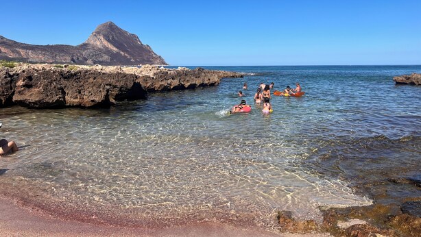 A beach with clear water, rocky shore, and a mountain in the background.