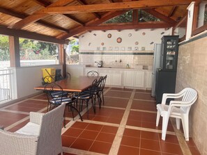 A covered patio with a wooden table and chairs, a refrigerator, and a tiled backsplash with decorative tiles. - Stunning Ocean Views in Macari - Apartment, Grill, and Air Conditioning (Macari)
