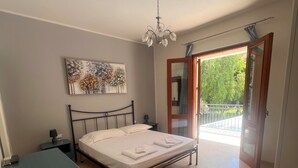 A bedroom with a bed, bedside tables, a dresser, a ceiling fan, a painting, and a balcony with a view of greenery.