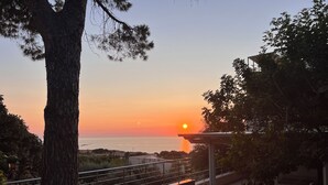 A sunset view from a balcony overlooking a body of water, with a railing and a pergola in the foreground.
