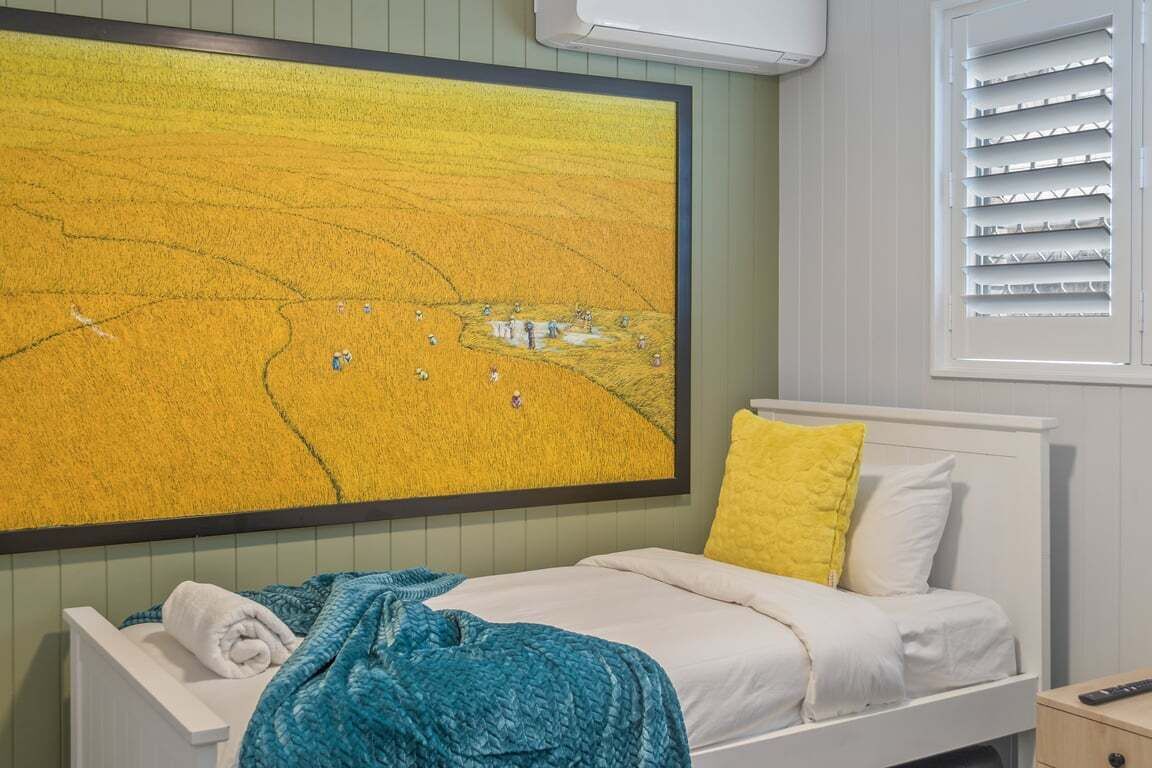 A bedroom with a large painting of a yellow field, a bed with white and yellow bedding, and a window with blinds.