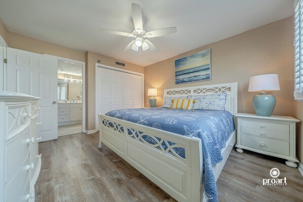 A bedroom with a bed, nightstand, dresser, ceiling fan, and artwork on the wall.