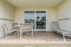 A balcony with a table and chairs, a lounge chair, and a sliding glass door leading to a view of palm trees.