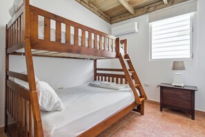 A bunk bed with a wooden frame, white bedding, a small bedside table with a lamp, and a window with blinds.