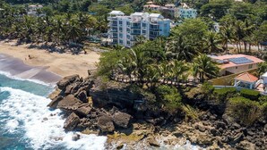 A coastal hotel with multiple buildings, a rocky shoreline, and palm trees. - Eco friendly beachfront oasis (Rincón)