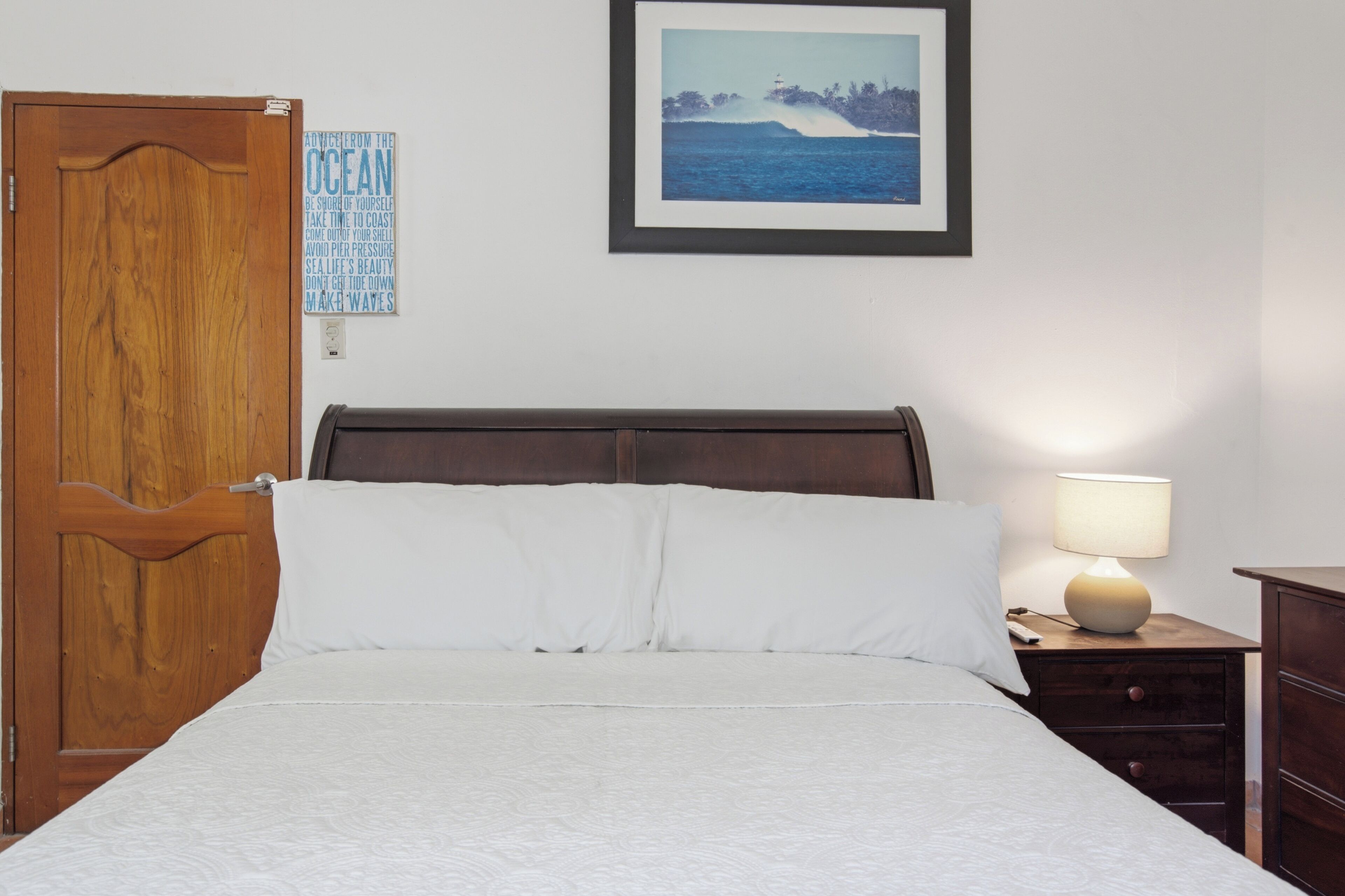 A bedroom with a wooden door, a framed picture of a beach scene, a bedside lamp, and a nightstand with a remote control.